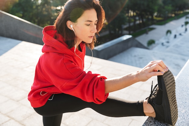 写真 イヤホンを着用して、屋外でストレッチ体操をしている美しい若いスポーツ女性