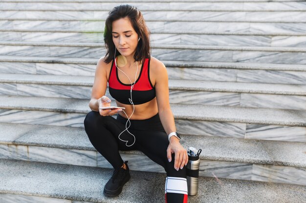 Beautiful young sportswoman holding mobile phone, listening to music with earphones, drinking water