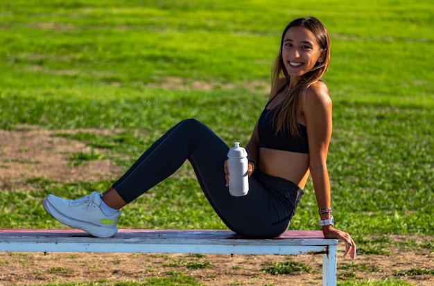 Beautiful young sporty girl tanned with long dark hair sitting on a wooden bench smiling happily