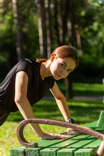 Foto bella giovane donna sportiva in maglietta nera, pantaloncini neri e scarpe da ginnastica rosa che si riscaldano esercitando i tricipiti e il torace facendo flessioni dalla panchina tra gli alberi prima di correre