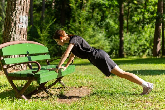 Beautiful young sporty woman in black tshirt black shorts and pink trainers warming up exercising triceps and chest dip doing pushups from bench among trees before running