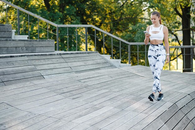 Beautiful young sporty woman walking with smartphone on the wooden pier or stairs in the park Smilling fitness girl smiling and looking to the phone