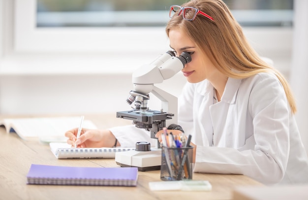 Фото Красивая молодая студентка-исследовательница смотрит в микроскоп и записывает записки.