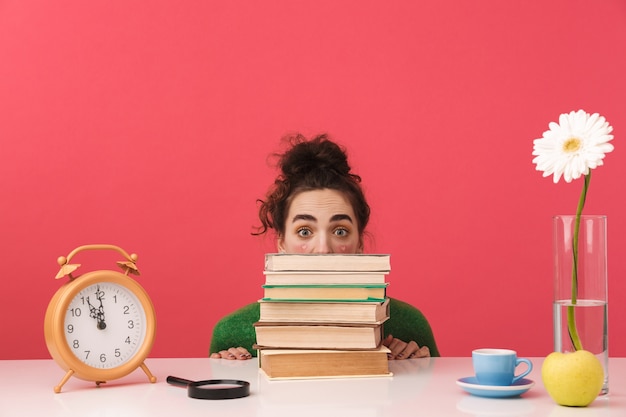 Photo beautiful young student girl sitting at the table isolated, studying with books, looking out