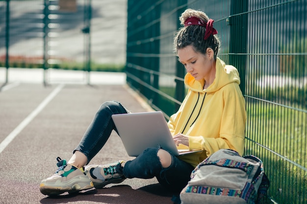 Foto bella giovane studentessa che indossa abiti casual e seduto sul campo sportivo con laptop moderno