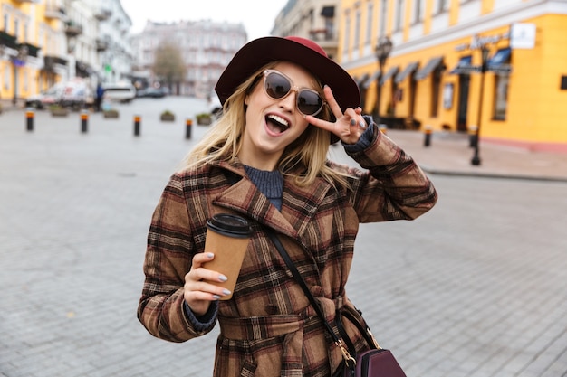Фото Красивая молодая стильная блондинка в пальто гуляет на свежем воздухе, держа чашку кофе на вынос