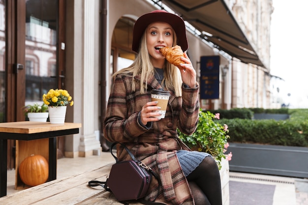 Bella giovane donna bionda alla moda che indossa un cappotto seduto all'aperto, mangiare croissant, bere caffè