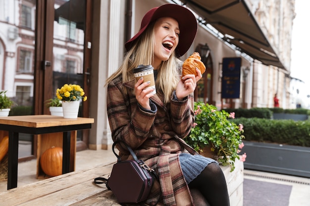 Beautiful young stylish blonde woman wearing a coat sitting outdoors, eating croissant, drinking coffee