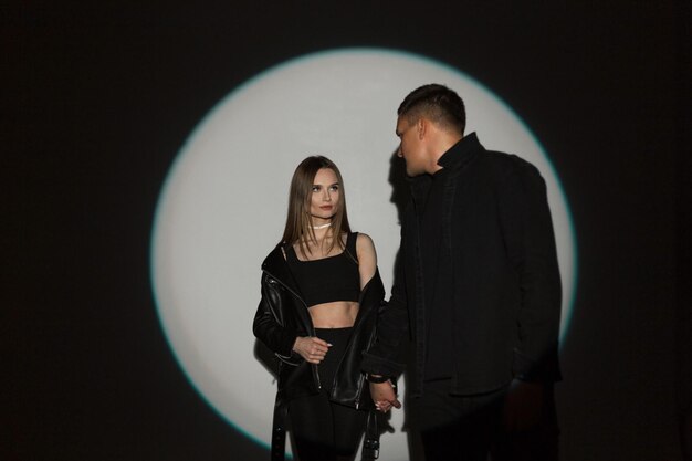 Beautiful young stylish couple girl and handsome man in fashion black rock casual clothes in the studio on a dark background with a hard circle light at a party