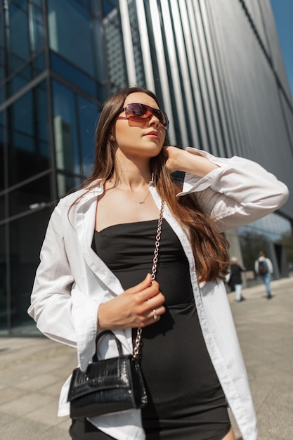 Фото Красивая молодая стильная элегантная модель девушки с солнечными очками в модном черном платье с белой рубашкой и сумочкой гуляет по городу
