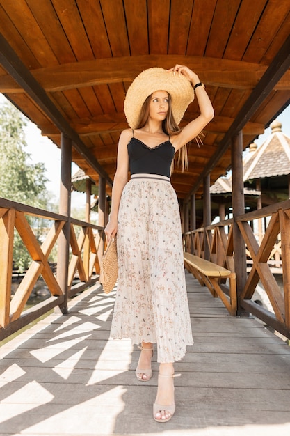 Beautiful young stylish girl in fashionable clothes with dress and lace shirt in a straw hat and wicker handbag walk on a wooden pier on vacation