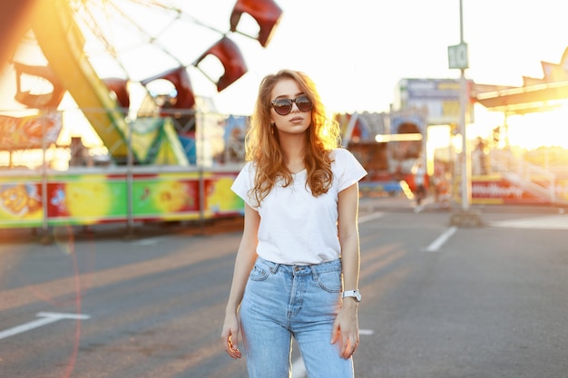Beautiful young stylish woman in sunglasses walking in the amusement park at sunset