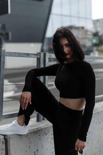 Photo beautiful young stylish woman with a messy bob hairstyle in fashionable black sportswear with white sneakers stands and poses near a modern building in the city