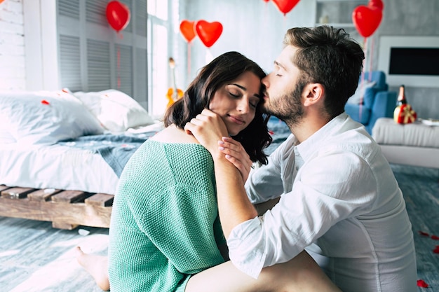 Beautiful young tender lovely couple are kissing and hugging each other while sitting on the floor of bedroom with romantic background. St Valentine's day. Anniversary. Date. Love concept