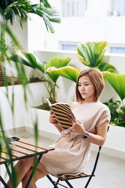 Bella giovane donna vietnamita concentrata sulla lettura di un libro interessante quando è seduto al tavolo del bar