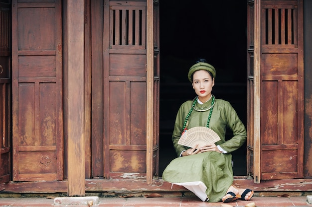 Beautiful young Vietnamese woman in traditional dress holding wooden fan when sitting on porch of old house