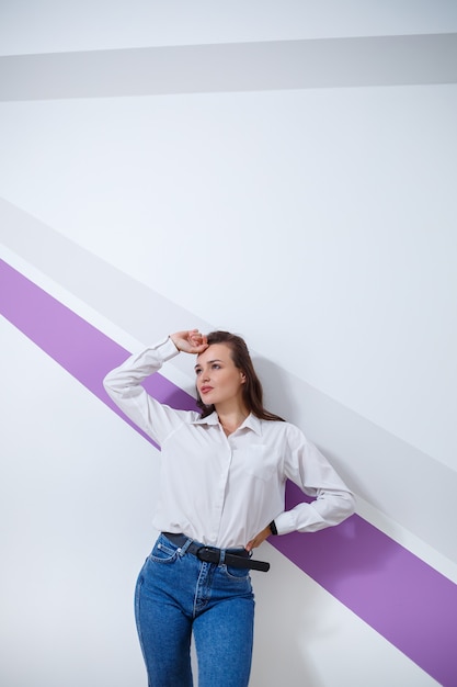 Beautiful young white-skinned girl dressed in a white shirt and blue jeans on a light background with a purple stripe