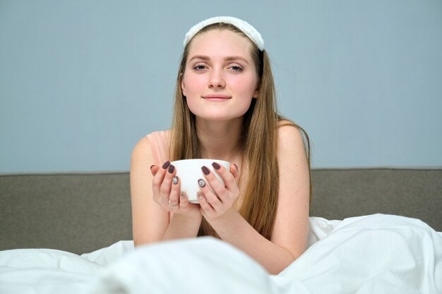 Beautiful young woman 20 years old blonde in bed with cup of tea, morning, white bed