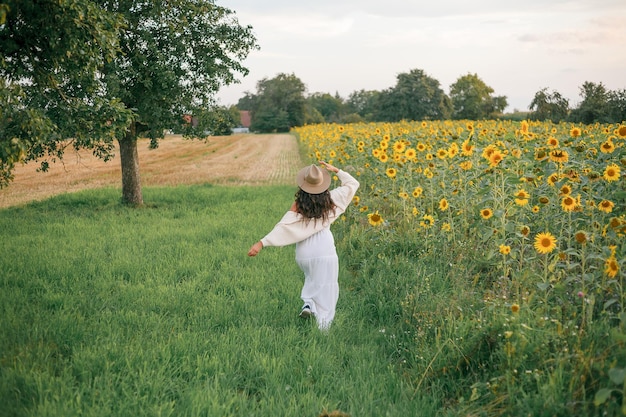 A beautiful young woman of 35 years old in a boho style hat a light white sundress runs at sunset in a sunflower field Summer evening Stylish model with curly dark hair is resting in the field