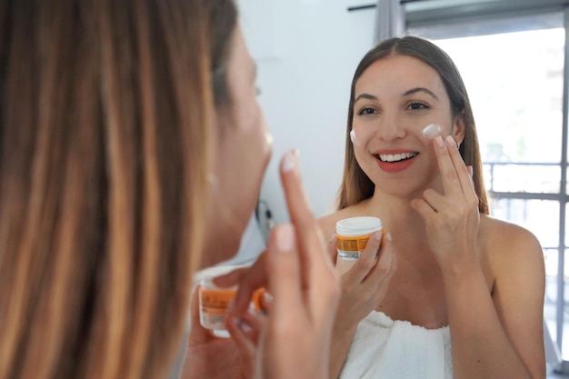 Photo beautiful young woman applying cream on her cheeks face moisturizing nourishing invigorating treatments enjoying relaxing time of skin care on mirror at home