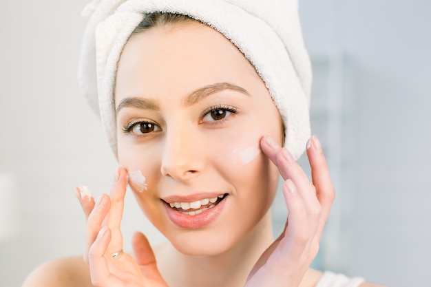 Beautiful young woman applying cream on her face in bathroom. Treatment for perfect skin concept