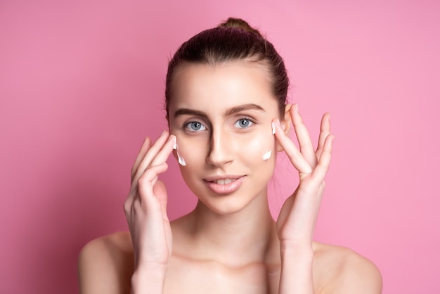 Beautiful young woman applying cream on her face on pink