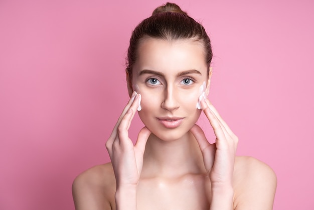 Beautiful young woman applying cream on her face on pink