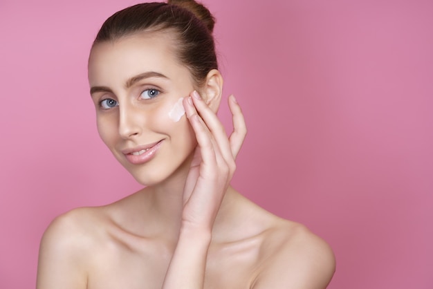 Beautiful young woman applying cream on her face on pink