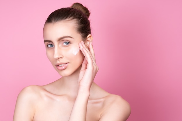 Beautiful young woman applying cream on her face on pink