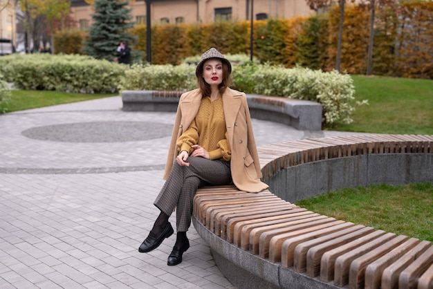 Beautiful young woman in an autumn beige coat and sunglasses sits on a flowerbed bench waiting for h...