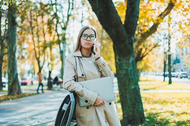 秋の公園で携帯電話で話しているリモートで作業しているラップトップと電話を使用して秋のコートを着た美しい若い女性