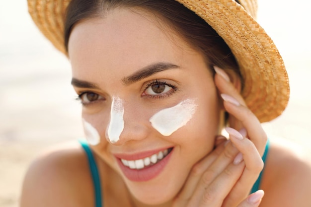 Photo beautiful young woman at beach applying sunscreen on face and looking at camera