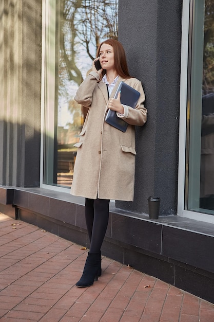 Beautiful young woman in beige coat with long hair holding laptop outdoors calling by smartphone