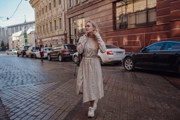 La bella giovane donna in un vestito lavorato a maglia beige cammina per le vie di mosca, tempo soleggiato della molla