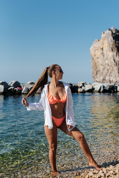 A beautiful young woman in a bikini walks along the ocean shore against the background of huge rocks on a sunny day Tourism and tourist trips Opening borders and vacation