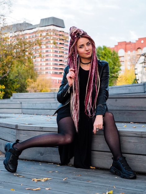 Beautiful young woman in black clothes with long pink dreadlocks sits on wooden steps and looks at the camera