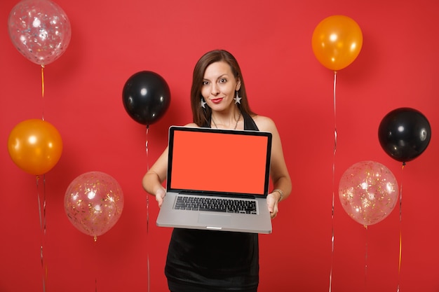 Beautiful young woman in black dress celebrating holding laptop pc computer with blank black empty screen on bright red background air balloons. Happy New Year, birthday mockup holiday party concept.