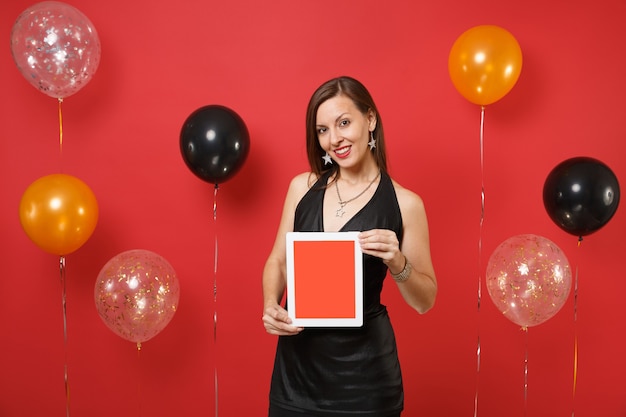 Beautiful young woman in black dress celebrating holding tablet pc computer with blank black empty screen on bright red background air balloons. Happy New Year, birthday mockup holiday party concept.