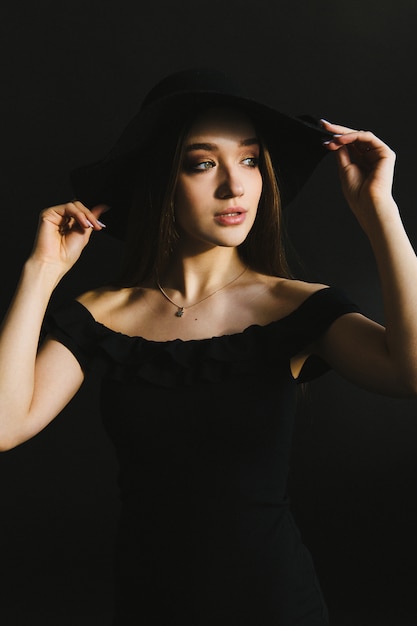 Beautiful young woman in black dress and hat on black background