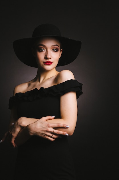 Beautiful young woman in black dress and hat on black background