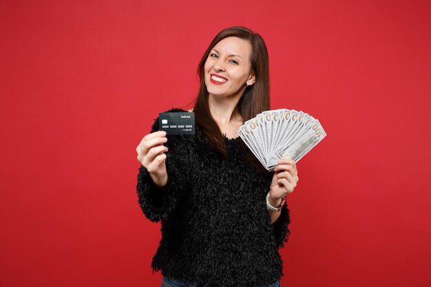 Beautiful young woman in black fur sweater hold credit bank card fan of money in dollar banknotes cash money isolated on red background. People sincere emotions, lifestyle concept. Mock up copy space.