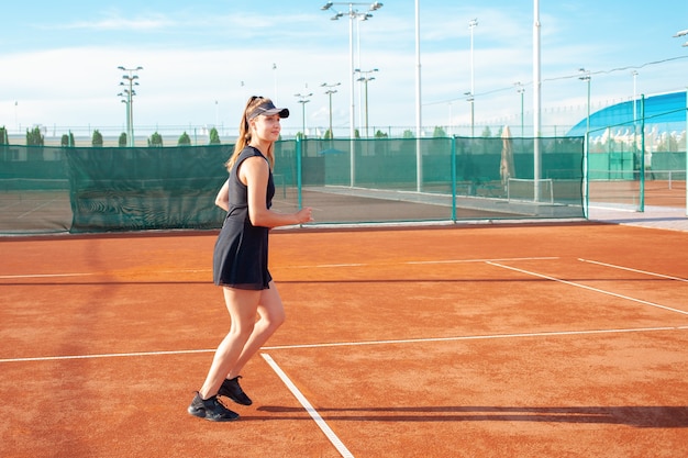 검은색 운동복을 입은 아름다운 젊은 여성이 테니스 코트에서 뛰어다닌다