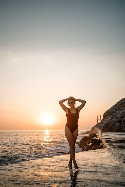 Красивая молодая женщина в черном купальнике одна на пляже у моря на закате. Взрослая стройная девушка отдыхает на берегу океана на закате. Концепция летних каникул. Выборочный фокус
