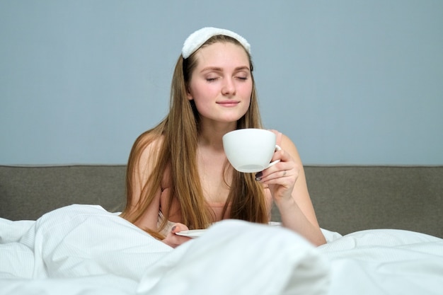 Bella giovane donna bionda a letto con una tazza di tè, mattina, letto bianco