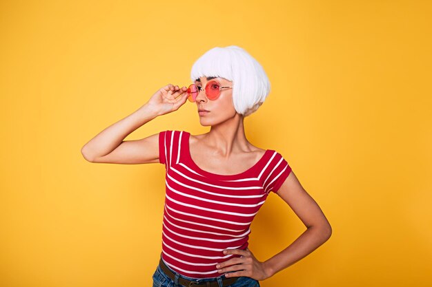 Beautiful young woman in blonde wig and pink sunglasses having fun on orange background fashion blonde posing