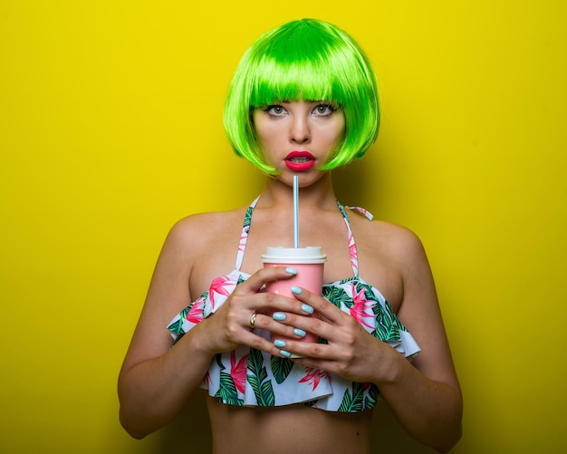 Beautiful young woman in a bright green wig under a quack and a swimsuit posing on a yellow background attractive girl with sensual lips flirts on the camera drinks a drink through a straw