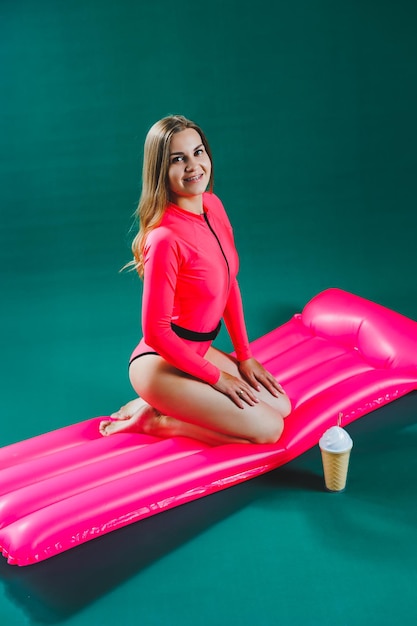 A beautiful young woman in a bright pink swimsuit sits on an inflatable pink mattress on a green background Summer rest