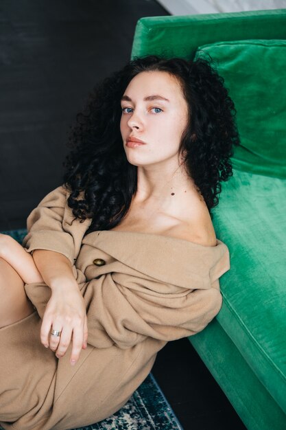 A beautiful young woman in a brown coat is sitting in a room on a green sofa. Interior.