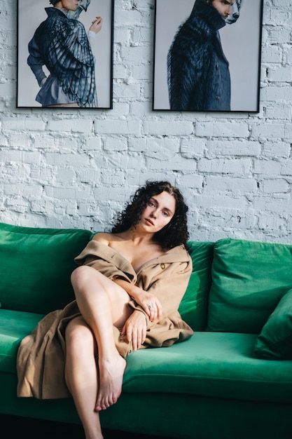 A beautiful young woman in a brown coat is sitting in a room on a green sofa. Interior.