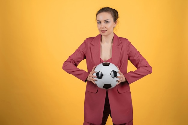 黄色の背景にポーズをとってサッカーボールを持つ美しい若い女性実業家
